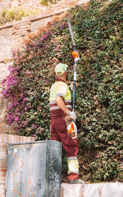 A gardener wearing a light green cap, a high-visibility vest with reflective stripes, and maroon work trousers is trimming a large, dense hedge with a long-reach garden pruning tool. The hedge is lush